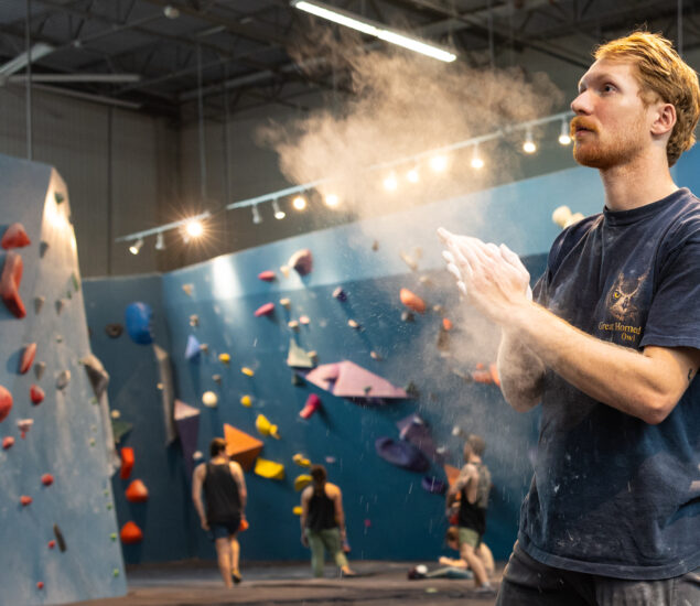 A man with red hair claps chalky hands in a brightly lit indoor bouldering gym, featuring colorful climbing holds, other climbers in the background, and K&N air filters in gyms for cleaner air.