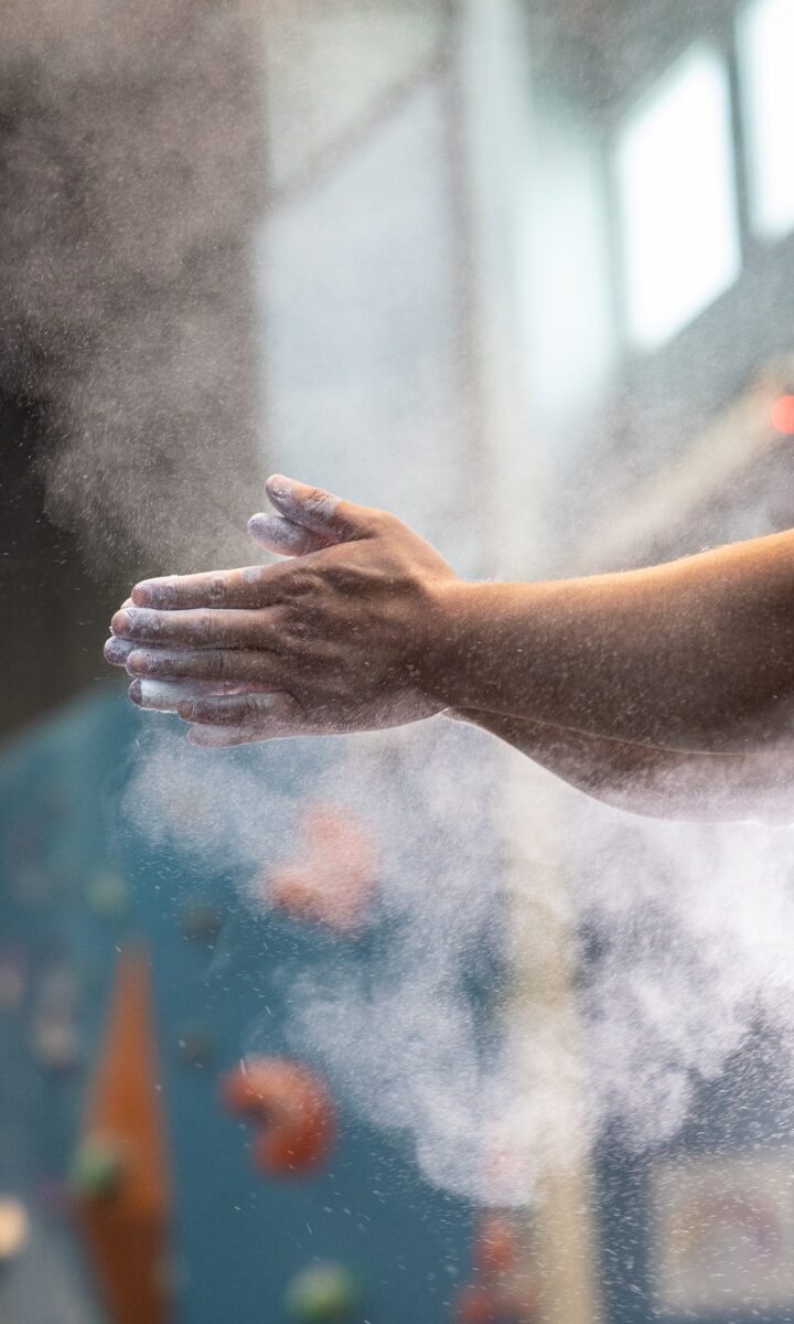 A close-up of a person clapping chalked hands, creating a cloud of chalk dust, with a blurred indoor climbing wall and K&N air filters in gyms helping keep the air clean in the background.