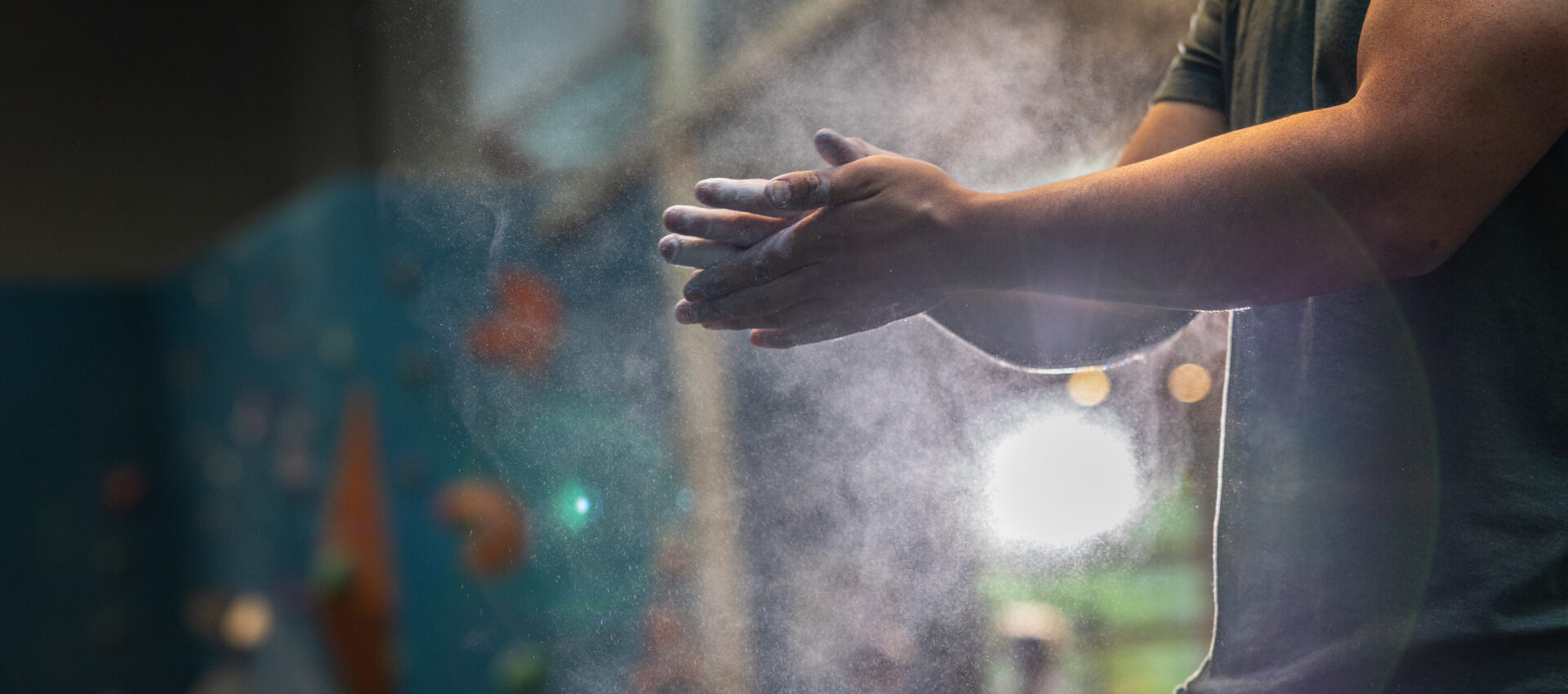 A person claps chalked hands together, creating a dust cloud, in an indoor climbing gym with a blurred bouldering wall and K&N air filters in gyms helping to keep the air clean in the background.