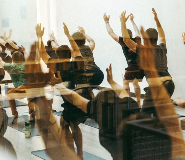 A group of people, on their First Time Visit, practicing yoga indoors and performing a lunge pose with arms raised. The image has a double exposure effect, creating overlapping, blurred silhouettes of the participants.