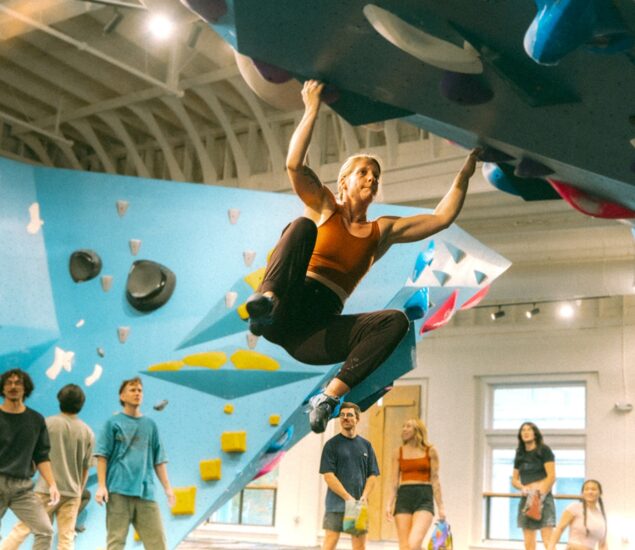 A woman in athletic wear climbs an overhanging indoor bouldering wall, gripping holds with her hands and feet. Several people, perhaps on their first time visit, stand below watching her. The gym has bright walls with various climbing routes.