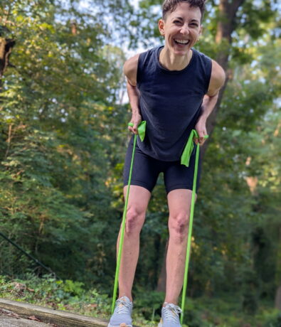 A person smiling and exercising outdoors in DC, leaning forward while pulling on green resistance bands. They are wearing a sleeveless black shirt, black shorts, and gray sneakers, with trees and greenery in the background. Perfect for personal training sessions.