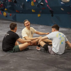 Three young men sit on the floor of Brooklyn Climbing Yoga and Fitness, laughing and talking together, with colorful climbing holds on the wall behind them.