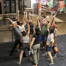 A group of people stand in a circle indoors at Brooklyn Climbing Yoga and Fitness, raising their arms and hands together enthusiastically as if celebrating or cheering in a vibrant recreational or gym space.