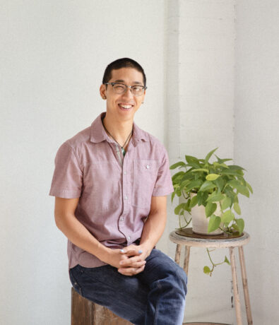 A person with short black hair and glasses sits smiling on a stool beside a green potted plant on a small round table, against the white upper walls of a bright, minimal Fremont room.