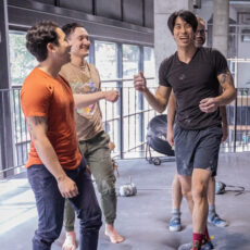 Four young men stand together indoors, laughing and smiling after a climbing session. One wears a black shirt and shorts, another an orange shirt and dark pants, a third a beige shirt with green pants, while a fourth is partially visible in the background.
