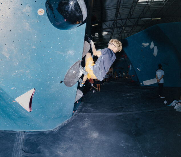 A person wearing a harness and yellow shorts climbs an indoor bouldering wall, gripping black holds while others watch in the background under bright lights—a perfect moment for a Youth Program landing page template.