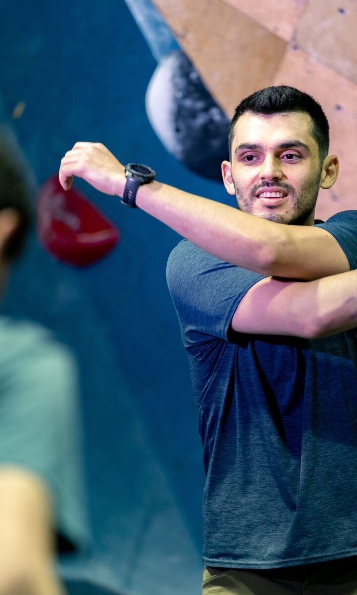 A man in a blue shirt stretches his arm across his chest in front of a DC climbing wall, with several Adult Team members standing nearby, slightly out of focus.