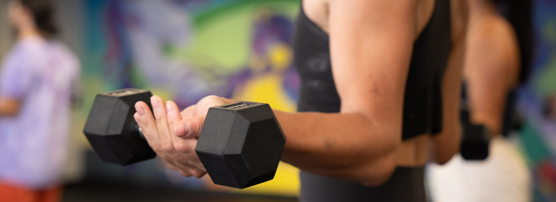 A person in athletic wear lifts a black dumbbell during a workout in a DC gym, with other people and colorful wall art in the blurred background—an inspiring scene of personal training in action.