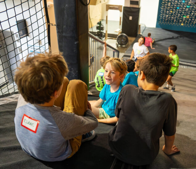 Several children sit and chat on a mat at DC Adventure Days indoor climbing gym, with colorful climbing walls and more kids in the background. One child in the foreground wears a name tag that reads ISAAC.