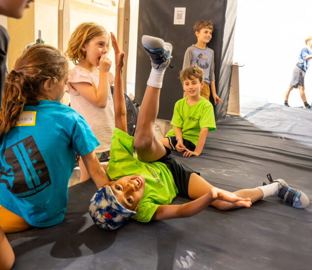 A group of children are gathered on a padded floor during Adventure Days, smiling and laughing. One child in a green shirt and hat lies playfully on his back with his legs in the air, while others watch and enjoy the moment.