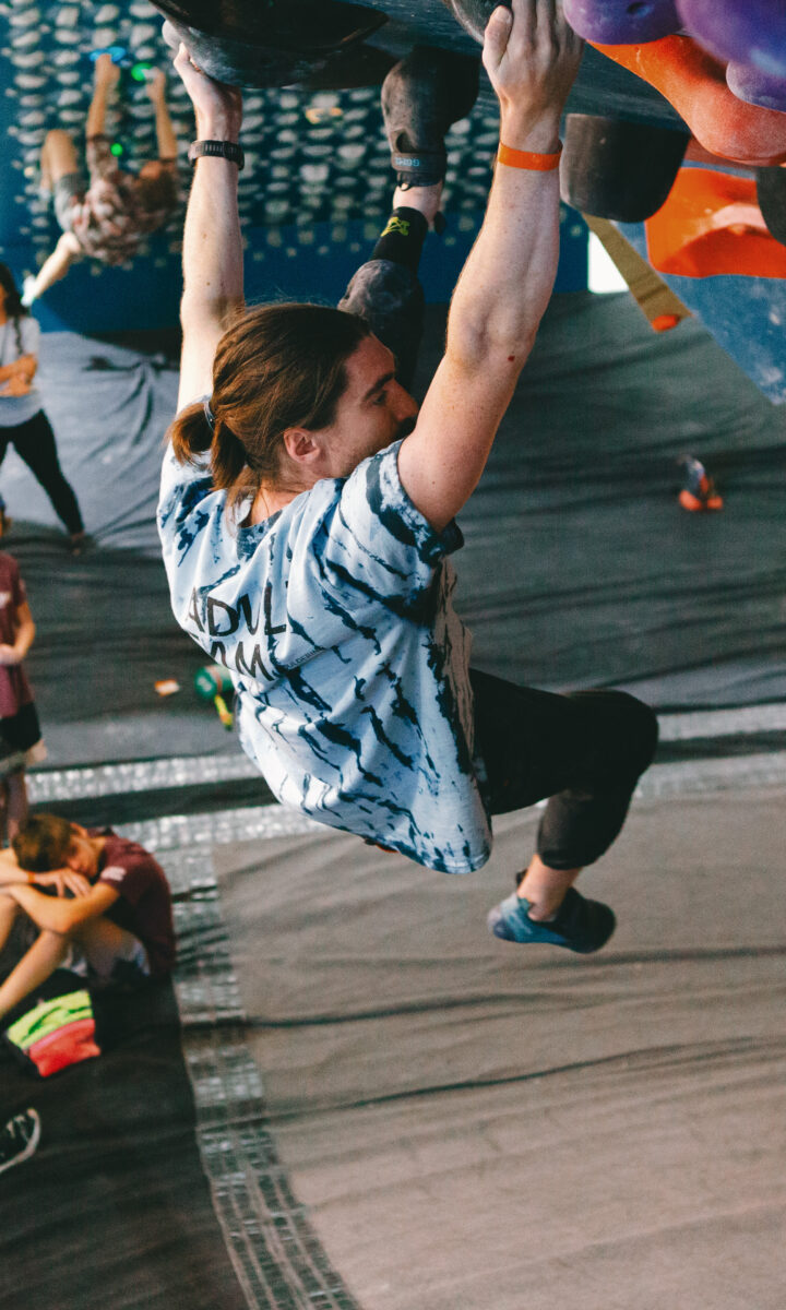 A member of the DC Adult Team with long hair tied back climbs an indoor bouldering wall while people watch and take notes below. The climber grips colorful holds on an overhanging section in the busy gym filled with spectators and participants.