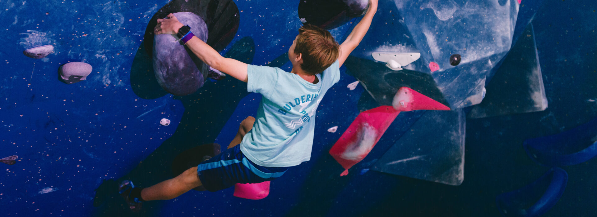 A person wearing a blue shirt climbs an indoor bouldering wall, gripping large purple holds on a blue surface—a great example of youth sports opportunities in DC.