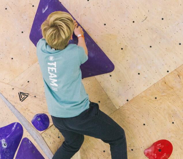 A person with short blond hair climbs an indoor bouldering wall in DC, gripping a large purple hold and standing on smaller red and purple holds. Wearing a blue TEAM shirt, they represent the spirit of Youth Teams in action.