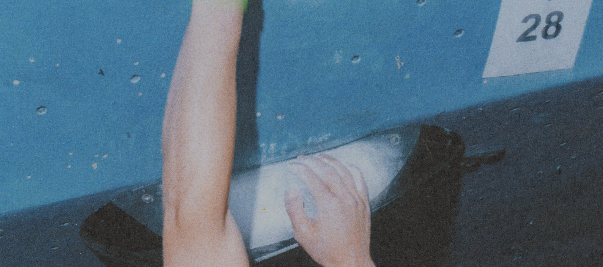 A person’s hands gripping climbing holds on a blue indoor climbing wall, with a white number 28 sign visible on the wall above the climber’s arm.