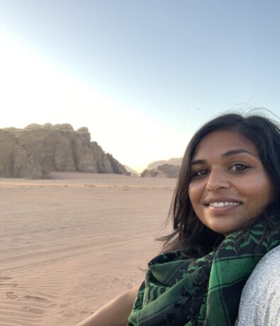 A woman with dark hair and a green scarf smiles at the camera while sitting on red desert sand after climbing, with rocky cliffs and a clear sky in the background.