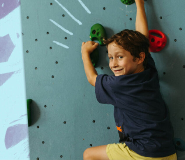 A young boy wearing a navy shirt and yellow shorts smiles while climbing an indoor rock wall with green holds at summer camp.