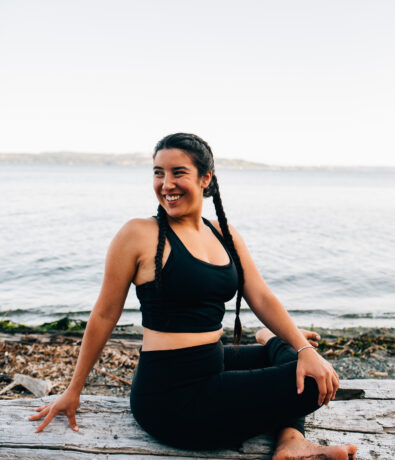A woman in black athletic wear sits on a log by the water near the University District Seattle, smiling while doing a seated yoga pose. The calm body of water and distant shoreline create a peaceful scene, perfect for post-climbing gym relaxation.
