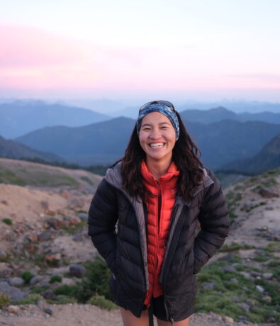 A woman stands smiling on a rocky mountain trail at sunset near the Upper Walls of Fremont, wearing a puffy jacket, red hoodie, shorts, and a blue headband, with mountain peaks and a pastel sky in the background.