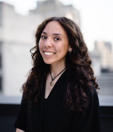 A young woman with long, curly brown hair smiles at the camera. She is wearing a black top and a necklace, standing outdoors near the University District Seattle, with a blurred cityscape—possibly close to a popular climbing gym—in the background.