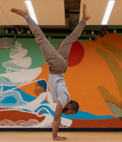 A man performs a handstand on a wooden floor, showcasing his climbing strength in front of a colorful mural featuring abstract shapes and a reclining figure.
