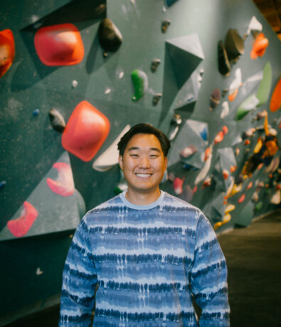 A person smiling and standing indoors in front of a climbing wall with various colorful handholds and grips.