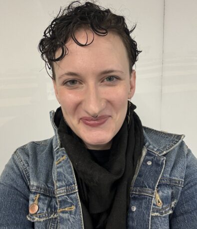 A person with short, curly dark hair smiles at the camera. They are wearing a denim jacket over a black scarf and shirt, with a white background behind them.