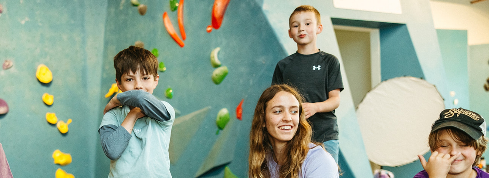 Four children smile and pose in an indoor rock climbing gym, with colorful climbing holds on a blue wall in the background. The cheerful, playful scene highlights the excitement of youth programs Boston has to offer.