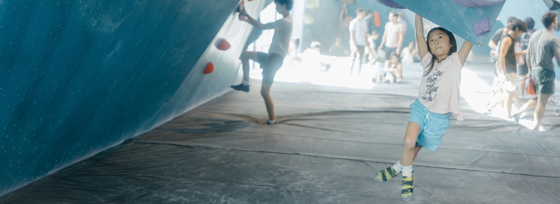 A young child hangs from a climbing wall in an indoor gym, wearing a t-shirt, blue shorts, and climbing shoes—a great example of youth activities. Other climbers and people are visible in the blurred background.