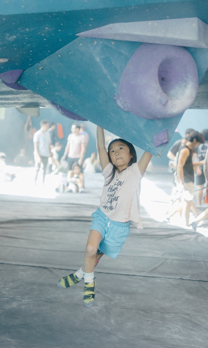 A young child wearing a t-shirt, blue shorts, and climbing shoes hangs from a large indoor climbing wall during one of the gym’s Youth Programs. Other people are visible in the background of the bustling climbing gym.