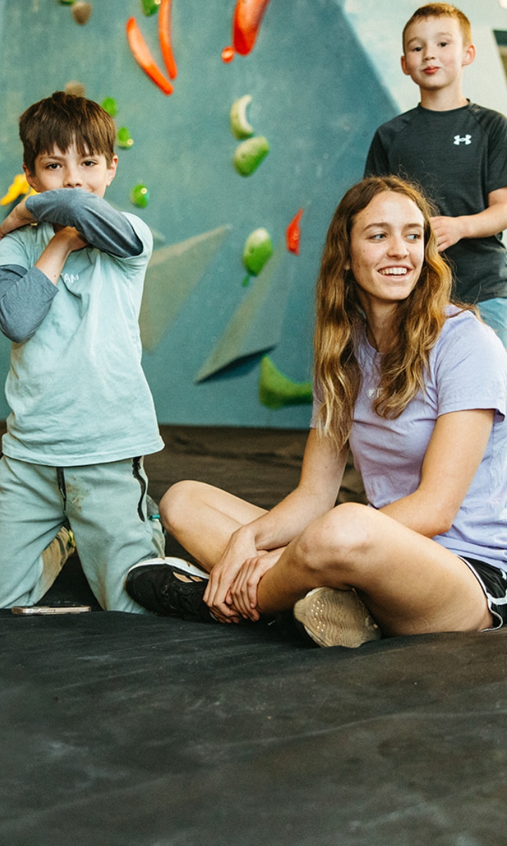 Three children are at an indoor climbing gym, part of youth programs Boston offers. One sits on the floor smiling, another stands with arms crossed, and a third stands behind them. Colorful climbing holds line the wall in the background.