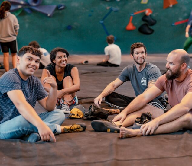 Four people sit on the floor in a climbing gym, smiling and chatting with climbing gear around them. Other climbers with Access Memberships and a colorful indoor climbing wall can be seen in the background.