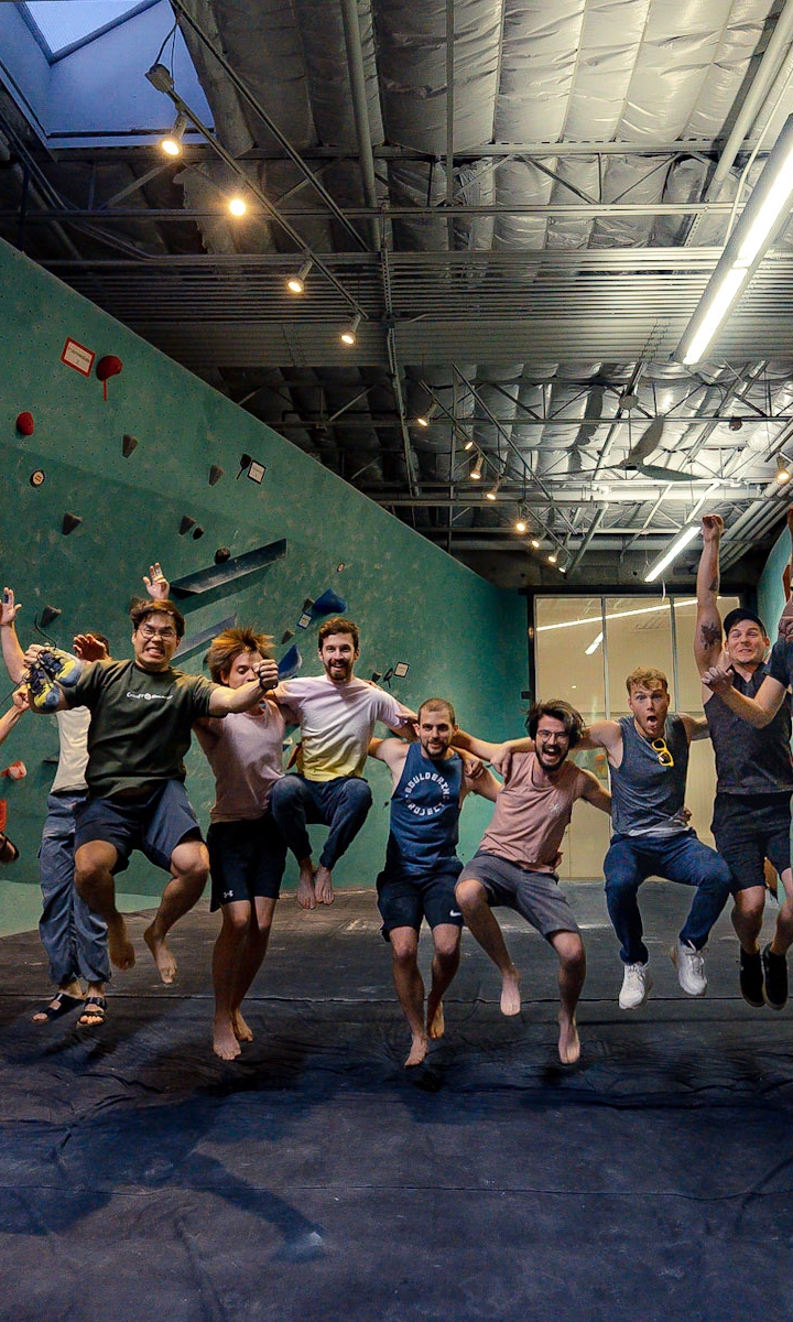 A group of eight people are joyfully jumping in the air inside an indoor climbing gym, celebrating their Access Memberships, with climbing walls and holds visible in the background.