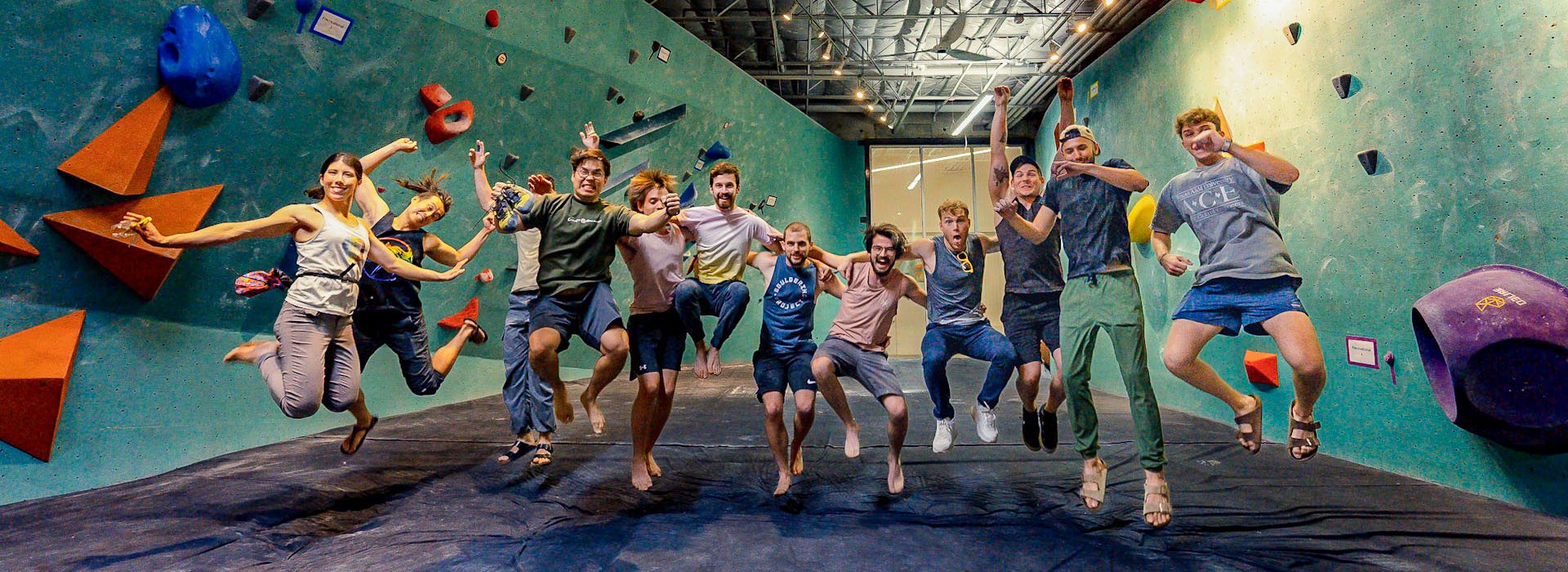 A group of twelve people with Access Memberships are jumping in the air and smiling inside an indoor rock climbing gym, with colorful climbing holds and padded flooring visible in the background.
