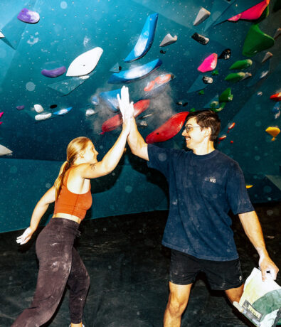 Two people in athletic wear high-five each other at a climbing gym in Seattle’s University District, chalk dust visible in the air, celebrating a successful climb against a colorful indoor wall.