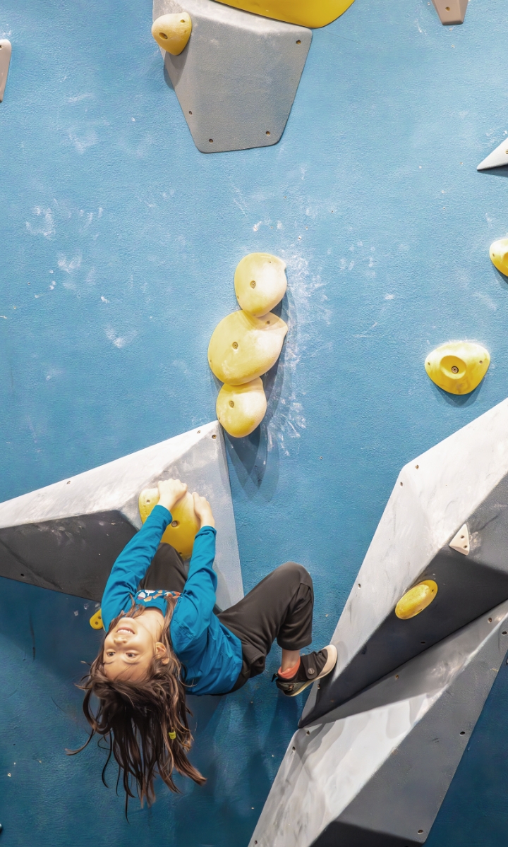 A young person with long hair climbs an indoor rock wall at a Brooklyn after school program, looking upward while gripping a yellow hold and balancing on angled surfaces with blue, gray, and yellow holds.