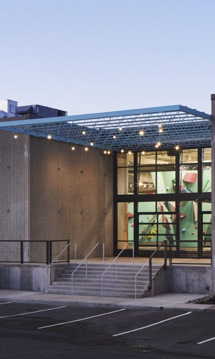 A modern Seattle building with large glass windows, a central staircase entrance, and a partially covered patio. The parking lot in front has several empty spaces, and the interior features colorful climbing walls. Contact Us for more details.