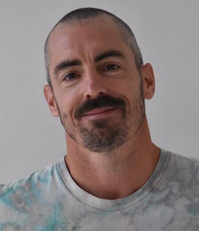 A man with closely-cropped hair, a mustache, and a short beard is smiling gently. He wears a light-colored tie-dye shirt and stands against a plain background, embodying the relaxed vibe of Brooklyn Climbing Yoga and Fitness.