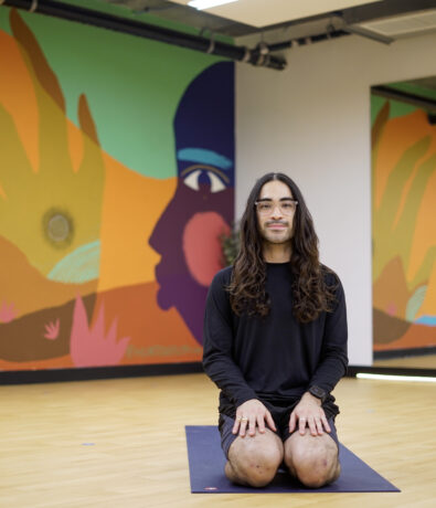 A person with long hair and glasses kneels on a yoga mat in a brightly colored room with abstract mural art inspired by climbing covering the wall behind them.