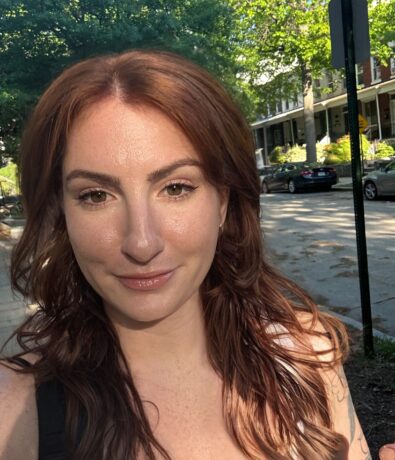 A woman with long, wavy auburn hair smiles slightly while taking a selfie outdoors on a sunny day, after climbing nearby trails; leafy trees, parked cars, and houses are visible in the background.