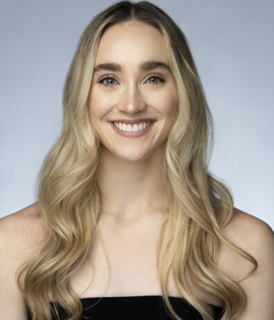 A woman with long, wavy blonde hair and light eyes smiles at the camera. She is wearing a black, strapless top, and the background is a plain, light gray—perfect for a profile photo near the climbing gym in University District Seattle.
