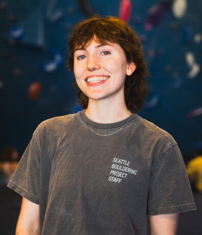 A smiling person with short brown hair wears a gray Seattle Bouldering Project Staff t-shirt and a necklace, standing in front of a blurred indoor climbing gym background, embodying the spirit of personal coaching in Seattle.