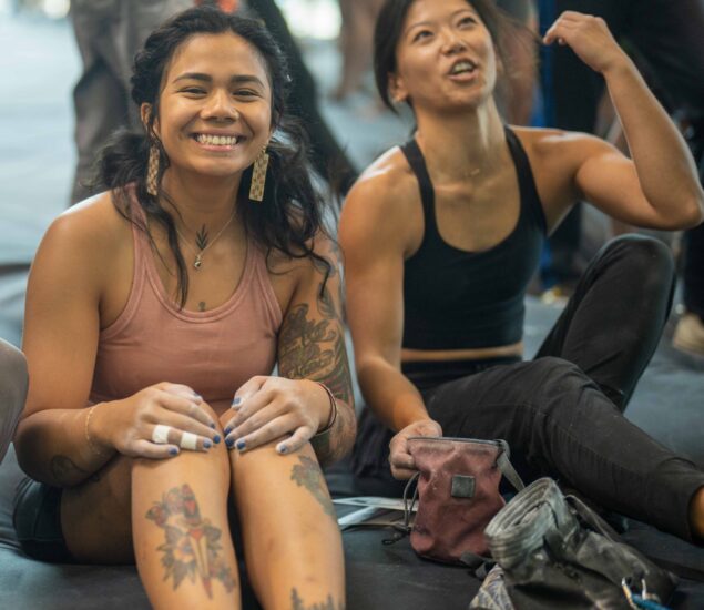 Two women in athletic clothing sit on the floor at Brooklyn Climbing Yoga and Fitness, smiling and relaxed after a workout. One has tattoos and wears a pink tank top; the other gestures with her arm. Gym bags are beside them.