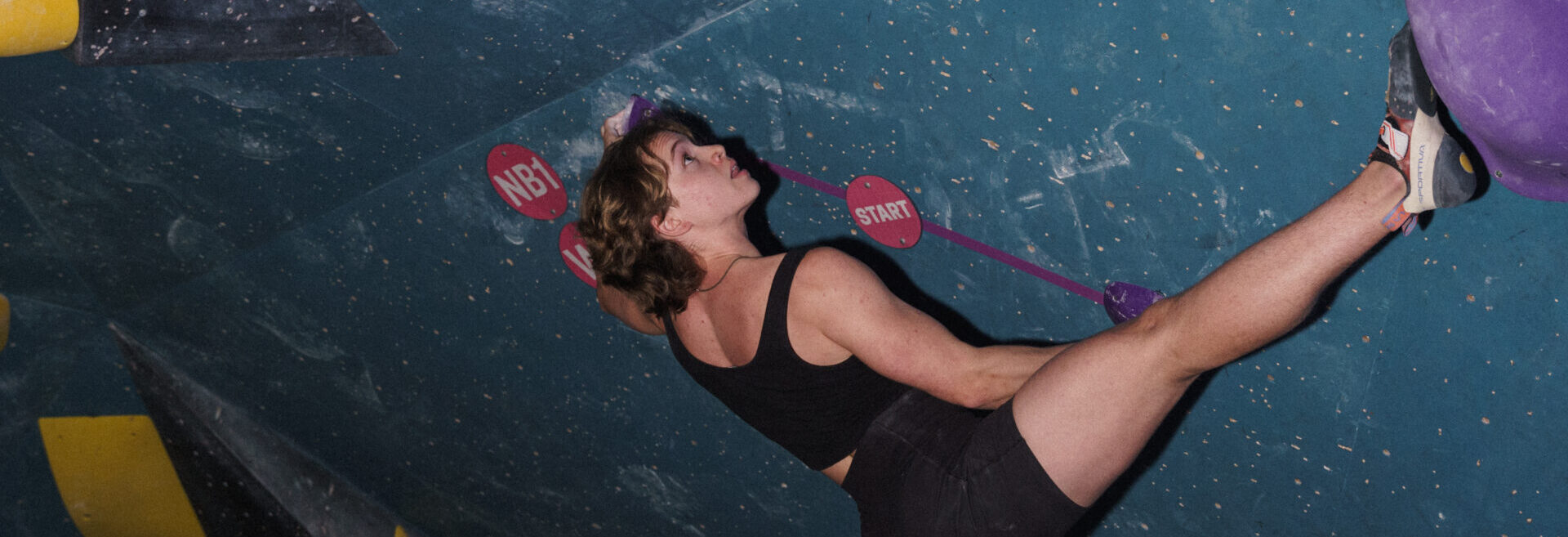 A person wearing a black tank top and shorts is climbing an indoor bouldering wall at Eckington, reaching up with one arm while their leg stretches out to a purple hold. The blue wall features climbing holds and chalk marks.