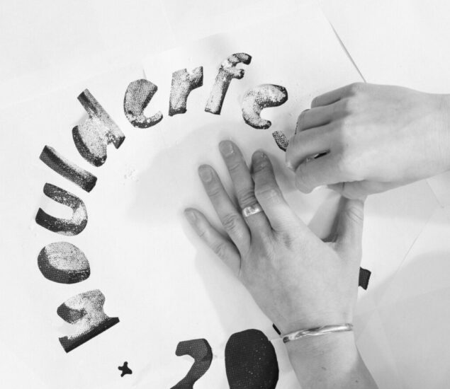 A person’s hands work on a stencil, revealing the word “Boulder” and the number “2021” on white paper. The black-and-white image hints at creative energy leading up to events like Boulderfest 2025, whether in Brooklyn or beyond.