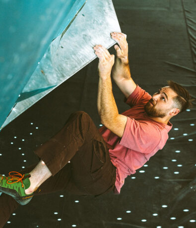 A man in a pink T-shirt and brown pants is bouldering indoors, gripping a large triangular climbing hold with both hands and looking upward in concentration.
