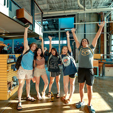 Five young adults stand indoors at an Austin climbing gym, arms raised and smiling, appearing cheerful and excited. Dressed casually in shorts and t-shirts, they enjoy the sunlight streaming into the spacious, modern room around them.
