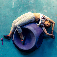 A person in striped pants and a short-sleeved top lies stretched out on a large purple climbing hold against a blue wall at an Austin Climbing Gym, with one arm extended above their head.