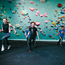 Four kids run indoors in front of a colorful wall at an Austin Climbing Gym, appearing energetic and happy as they play together.