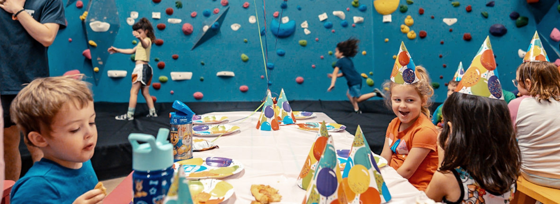 Children wearing party hats sit around a table with food and drinks at an indoor birthday party. In the background, kids climb a colorful rock climbing wall, making Birthday Parties here lively and festive.